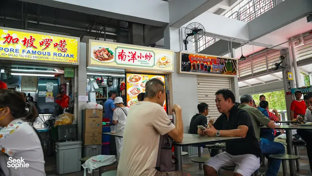 Singapore hawker centre tables where diners chat and eat by a busy stall counter beneath rojak menu signs — Singapore hidden gems