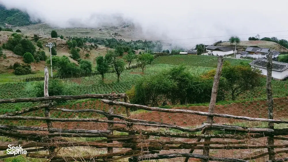 Misty countryside near Lijiang with a fenced foreground and a rural village of white houses as clouds roll through the valley