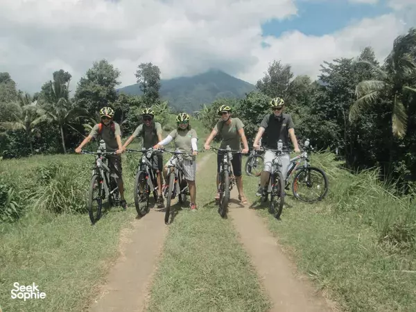 Group on Green Bikes Bali electric bikes on a countryside lane through Jatiluwih rice terraces, Bali, with mountains under clouds