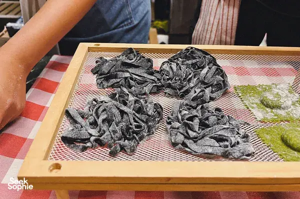 Sliced black pasta dough pieces drying on a mesh tray with green herbs in the foreground during workshops in Singapore