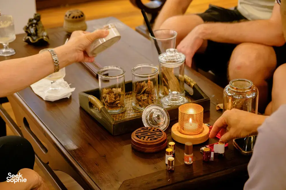 Hands selecting small glass vials on a tasting tray at the Rare Agarwood Tour in Hanoi, with scent samples and cups arranged nearby