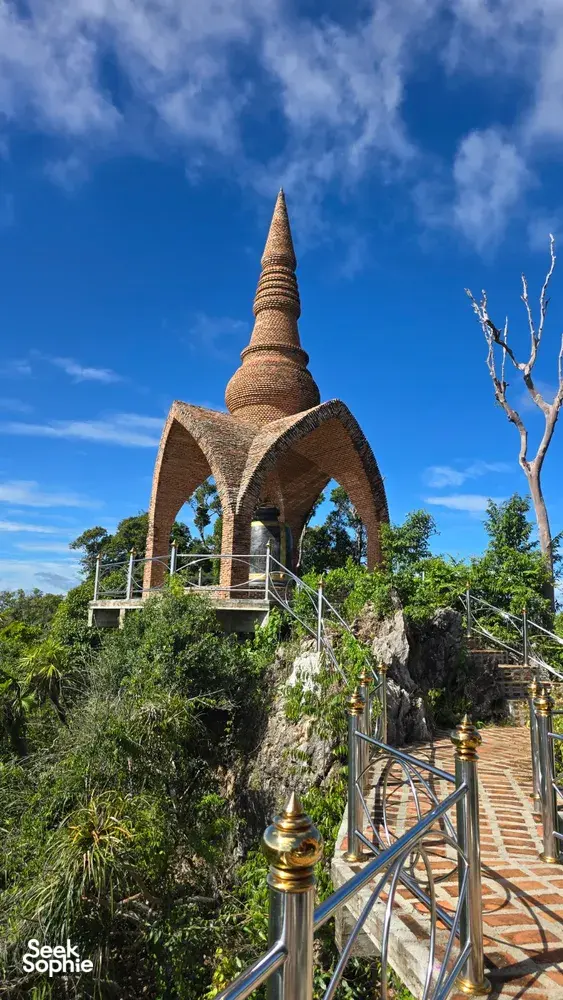 Bronze pagoda tower rising above a garden walkway with metal railings under a blue sky in Khao Sok National Park