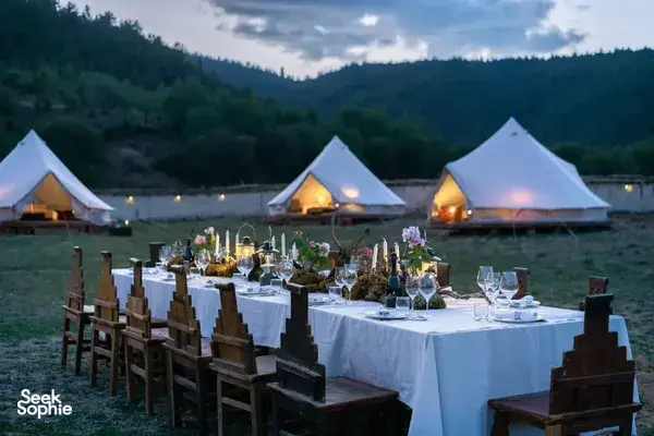 Candlelit outdoor dining table set with wine and starters in front of glowing white tents overlooking valley hills in Shangri-La
