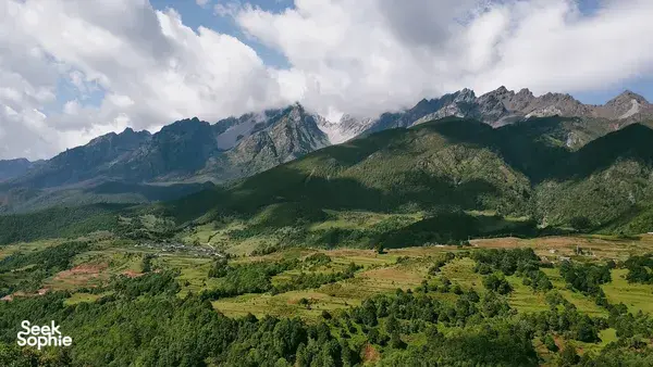 Lush green landscape around steep peaks near Lijiang, viewed from above with scattered clouds drifting over the mountains