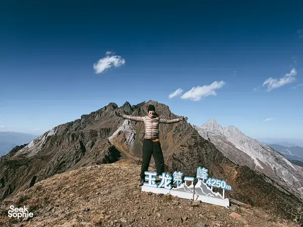 Hiker posing by a stone sign reading Jade Dragon 1st Peak, 4250m, during a guided summit hike in Lijiang
