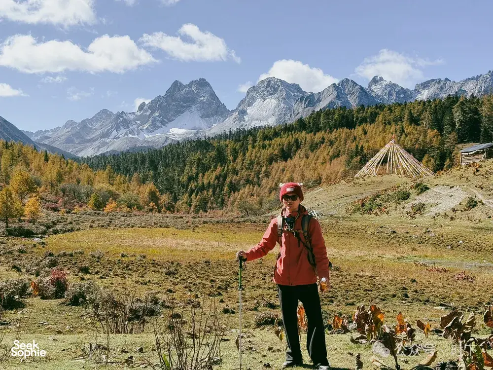 Hiker with trekking poles standing in a grassy alpine valley with a small tent as snow-capped mountains loom in Shangri-La