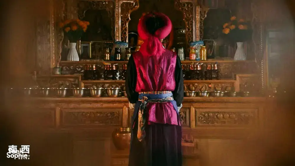 Back view of a person in ceremonial red robes and tall hat facing a shrine of brass bowls at a Khampa Caravan homestay in Shangri-La