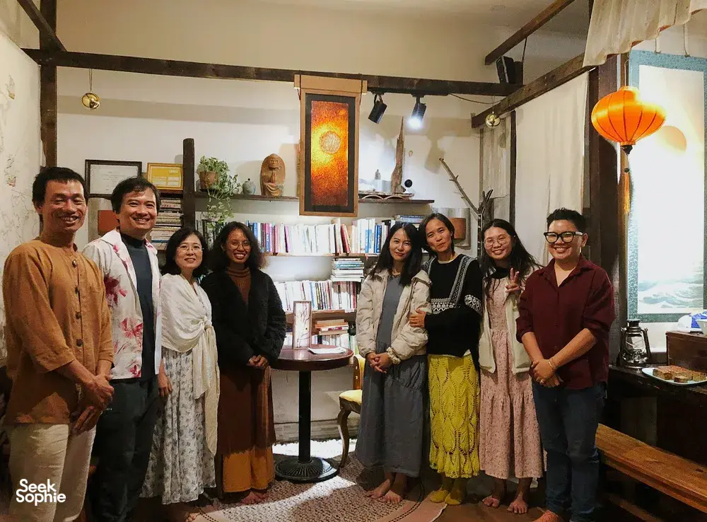 Sekelompok orang berkumpul di ruang perpustakaan yang nyaman di Chan Dat Living Room, Hoi An, dikelilingi rak, lampu lentera, dan buku saat menikmati pengalaman budaya yang mendalam