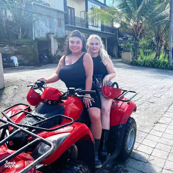 Two riders posing on red ATV quad bikes in Bali, sitting close together with palm plants and a building behind