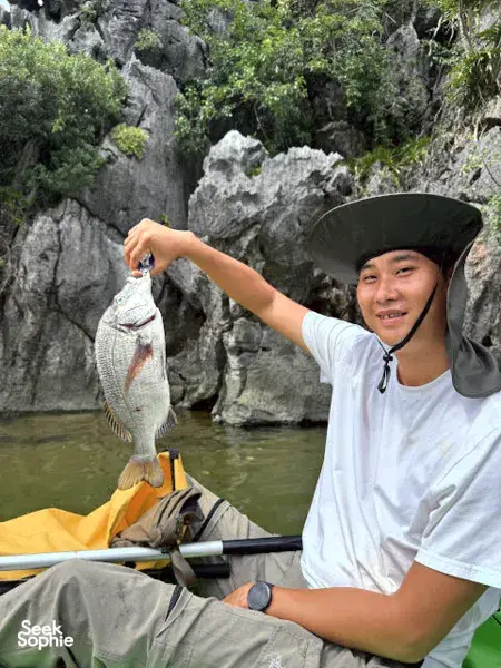 Tien Anh, Cat Ba local fishing enthusiast, holds up a freshly caught fish from a small boat near limestone cliffs