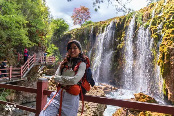 Hiker posing by a multi-tier waterfall in Shangri-La, standing on a wooden platform with red handrails and trekking gear