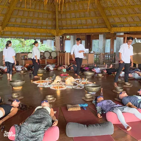 Sound-bath meditation in a wooden yoga hall with guests reclining on mats while singing bowls are played, Canggu