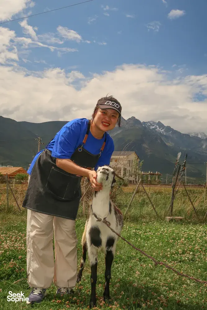 Seeker Yunnan Coffee Estate host smiling while cradling a black-and-white goat on the flowered coffee farm in Lijiang