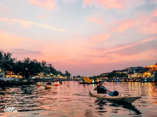 Kayaker gliding through Hoi An waterways at dusk, with palm trees and rows of lit riverside buildings stretching into the distance