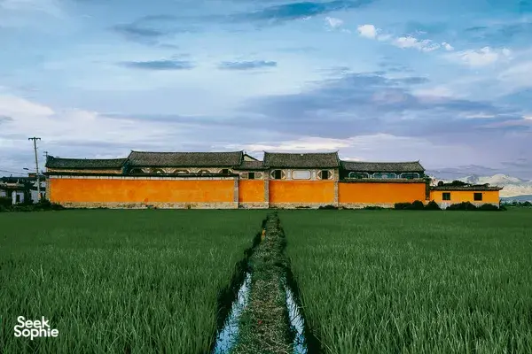 Orange-and-green Bai-style heritage buildings along the field in Xizhou, Dali, with mountains on the horizon