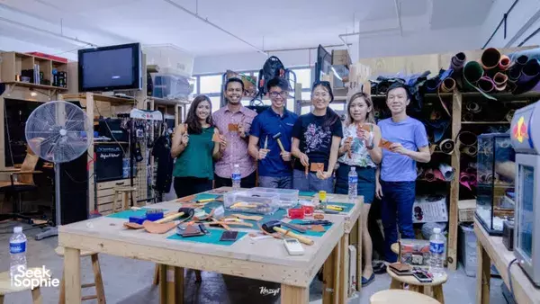Group posing with leather clutch pieces on a shared workbench inside a Singapore leather workshop — workshops in Singapore