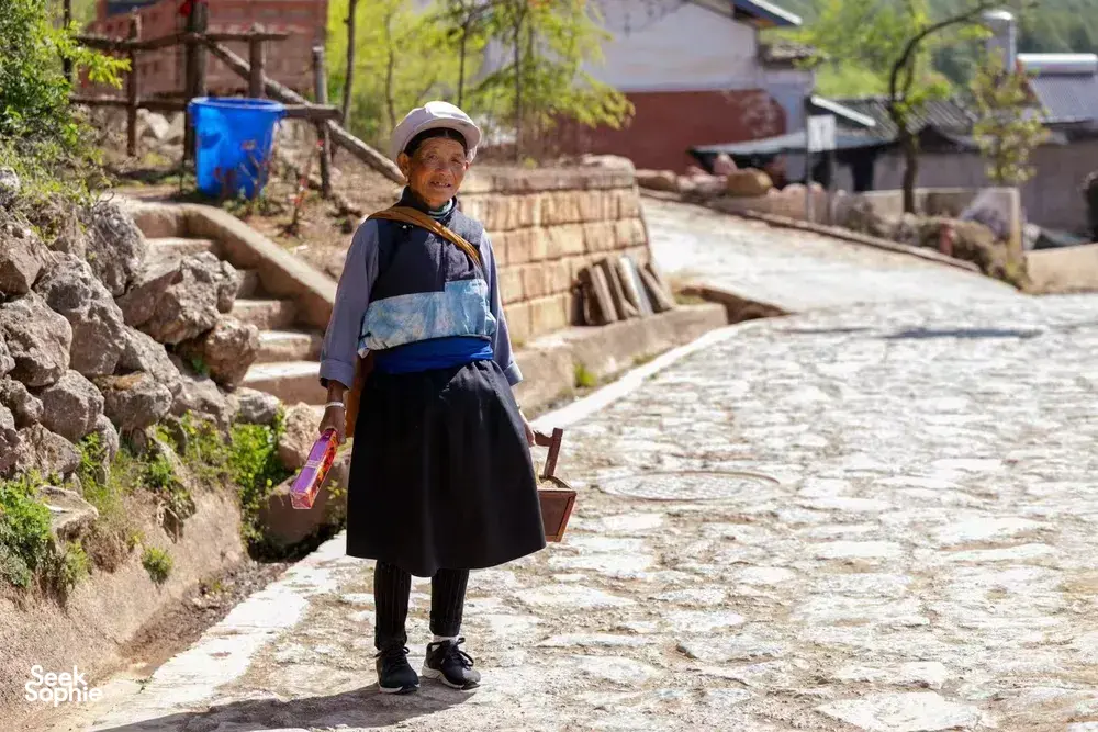 A porter carrying a large crate on a narrow stone lane in rural China, with village houses and trees in view