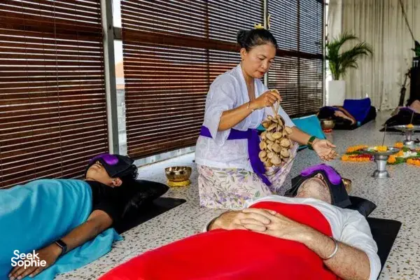 Practitioner holding wooden chimes beside a guest lying on a mat, with brass singing bowls set out for sound healing in Canggu