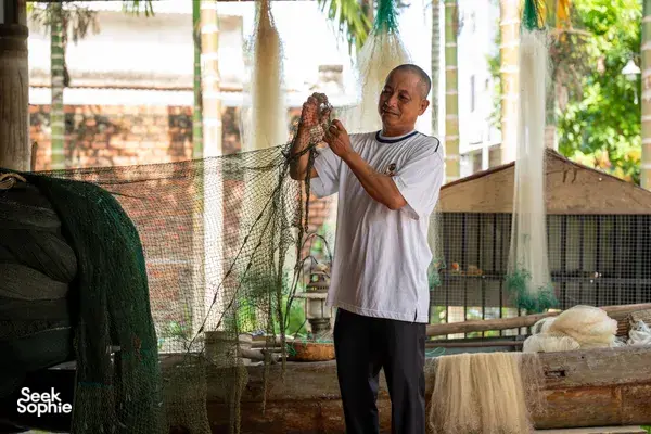Elderly man holding a gathered netting panel beside baskets of fishing gear at a riverside home in Hoi An