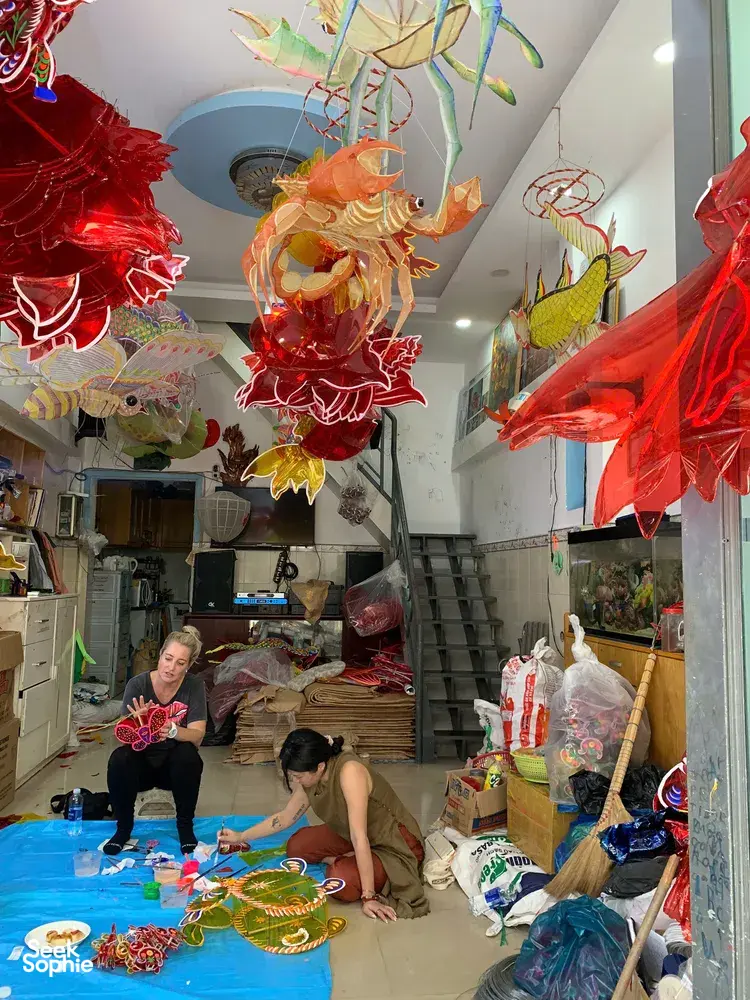 Traditional lantern workshop in Saigon with red and gold paper lanterns hanging overhead as makers work at tables in the shop