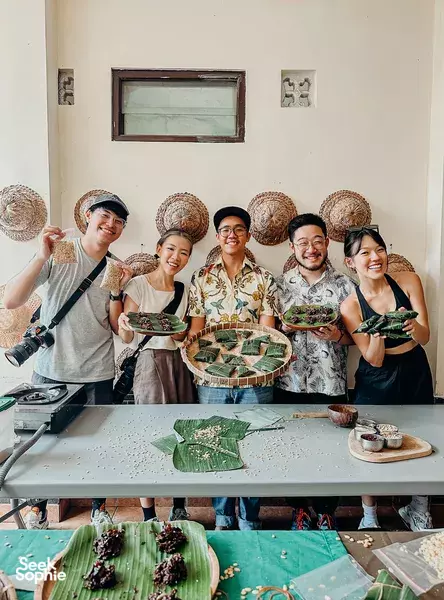 Group posing with banana-leaf parcels and leafy wraps at a cooking table during a Bali cooking class in a simple studio space