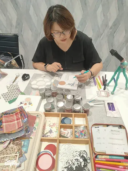 A participant adds details to her vision board for gemstones in Singapore, surrounded by collage templates, pens and jars; workshops in Singapore