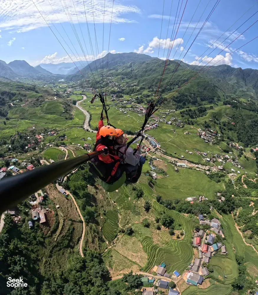 Tandem paragliding from behind with a helmeted rider guiding below the canopy over a valley of terraces and winding roads in Sapa