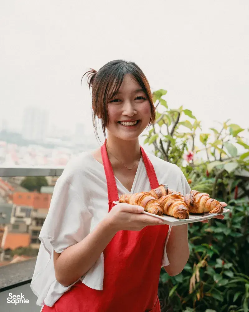 Smiling participant holding a tray of croissants on a balcony with city views, during a French Cooking Lab class in Singapore