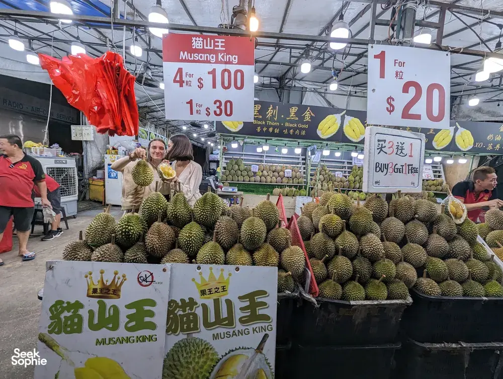 Durian stall in a Singapore market hall with piles of Musang King fruit and price signs, on a Geylang night walk