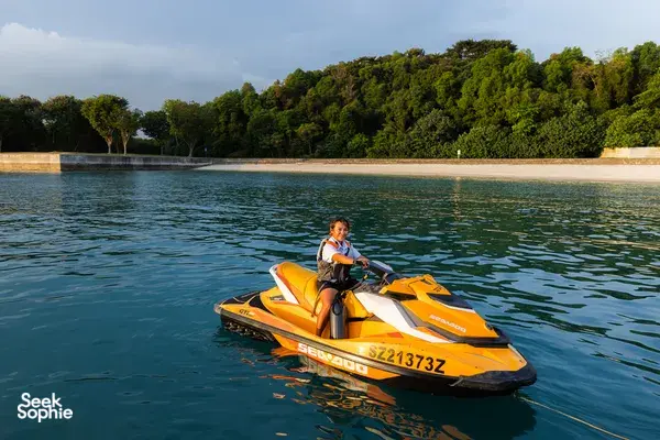 Orange jet ski speeding across calm waters near Singapore’s forested shoreline, with a rider wearing a life jacket
