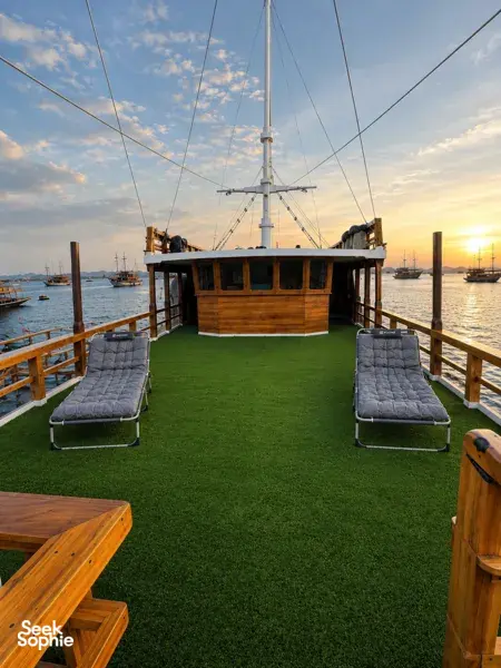 Sunset deck on a wooden phinisi with green carpet, padded loungers, and anchored masts overlooking the harbour in Labuan Bajo