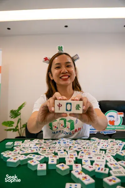 MahjongLah host smiling and pointing at mahjong tiles from a green table covered in hands of tiles in Singapore — team building Singapore