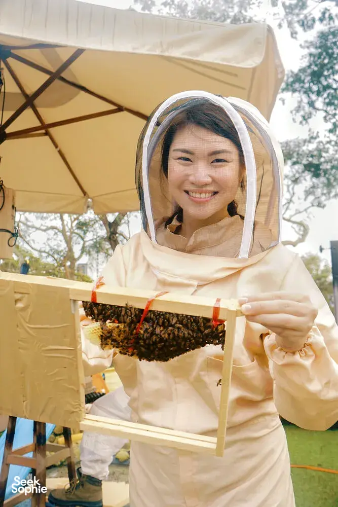 Smiling beekeeper in protective veil holding a honeycomb frame under a canopy tent during beekeeping in Singapore