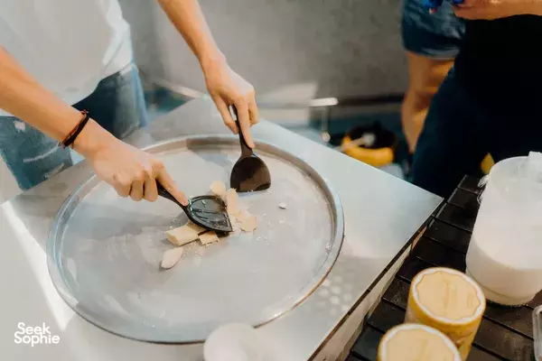 Hands scooping and spreading ice-cream base onto a chilled tray with tubs on the counter during ice-cream making workshops in Singapore