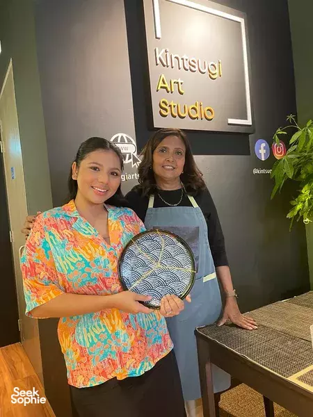 Two people in aprons holding a gold-flecked repaired ceramic plate at Kintsugi Art Studio, learning kintsugi during workshops in Singapore