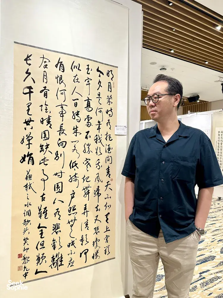Chong Zheng Calligraphy host in Singapore standing beside a large Chinese calligraphy scroll display at an indoor hall