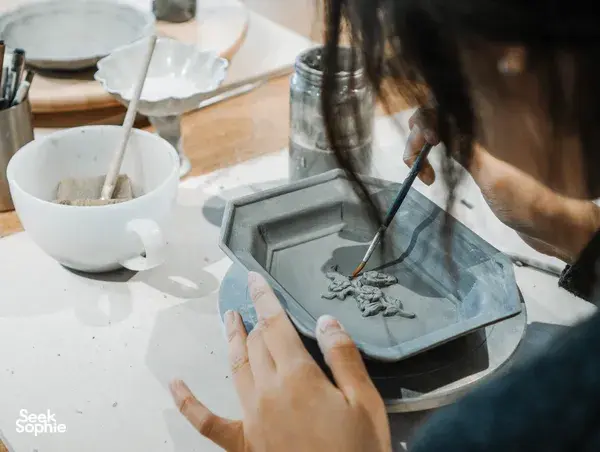 Hands using a brush to paint and position small ceramic pieces on a tray during a Singapore pottery workshop at the worktable