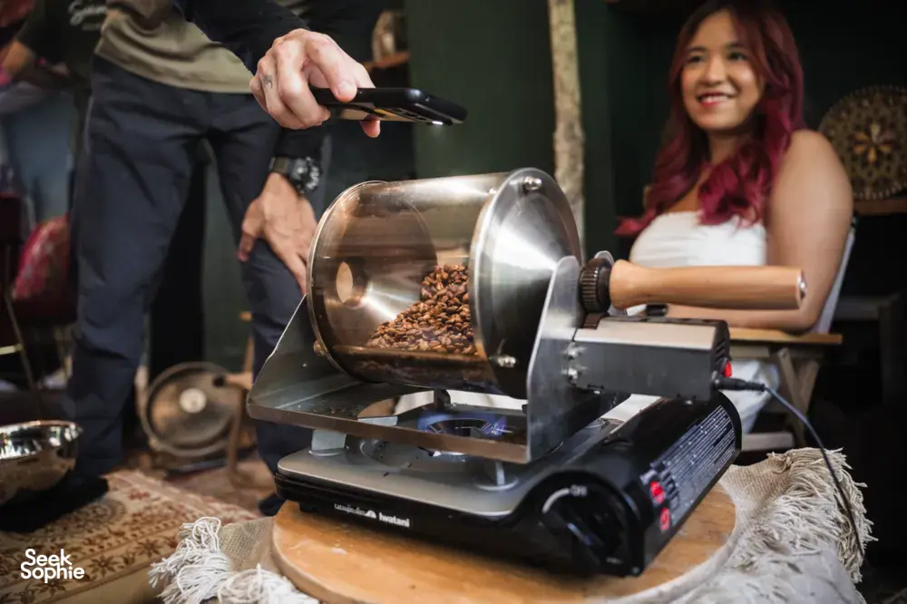 Beans being tipped into an electric coffee roaster as a woman smiles in the background during hands-on coffee roasting workshops in Singapore