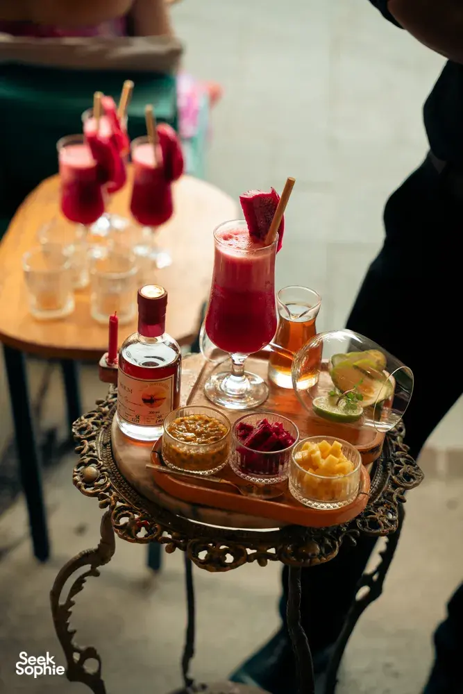 Koktel merah dalam gelas berkaki dengan hiasan tembikai di atas dulang hidangan hiasan, bersama botol spirit dan sajian pembuka, Hoi An