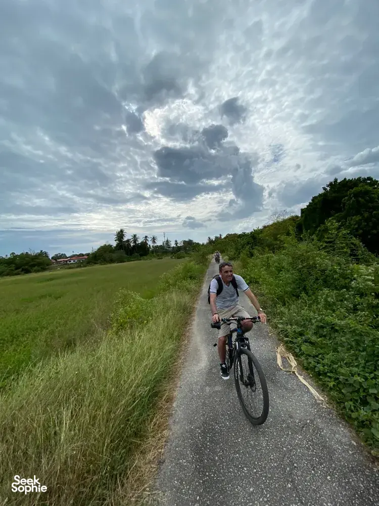 Cyclist pedals along a narrow rural road in Langkawi, past tall grass and hedges under bright, dramatic clouds