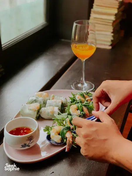 Hands assembling fresh spring rolls with herbs and dipping sauce at Chan Dat Living Room, Hoi An, with orange drink
