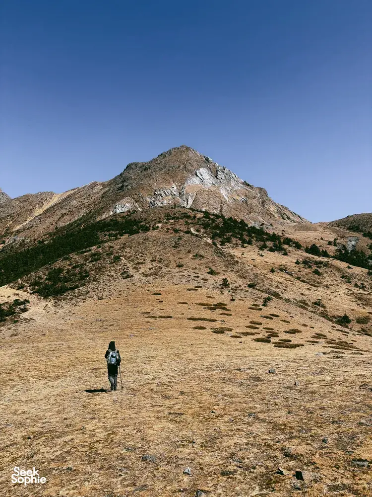 Solo hiker crossing a barren alpine slope toward a jagged peak on a guided hike of Jade Dragon Snow Mountain in Lijiang