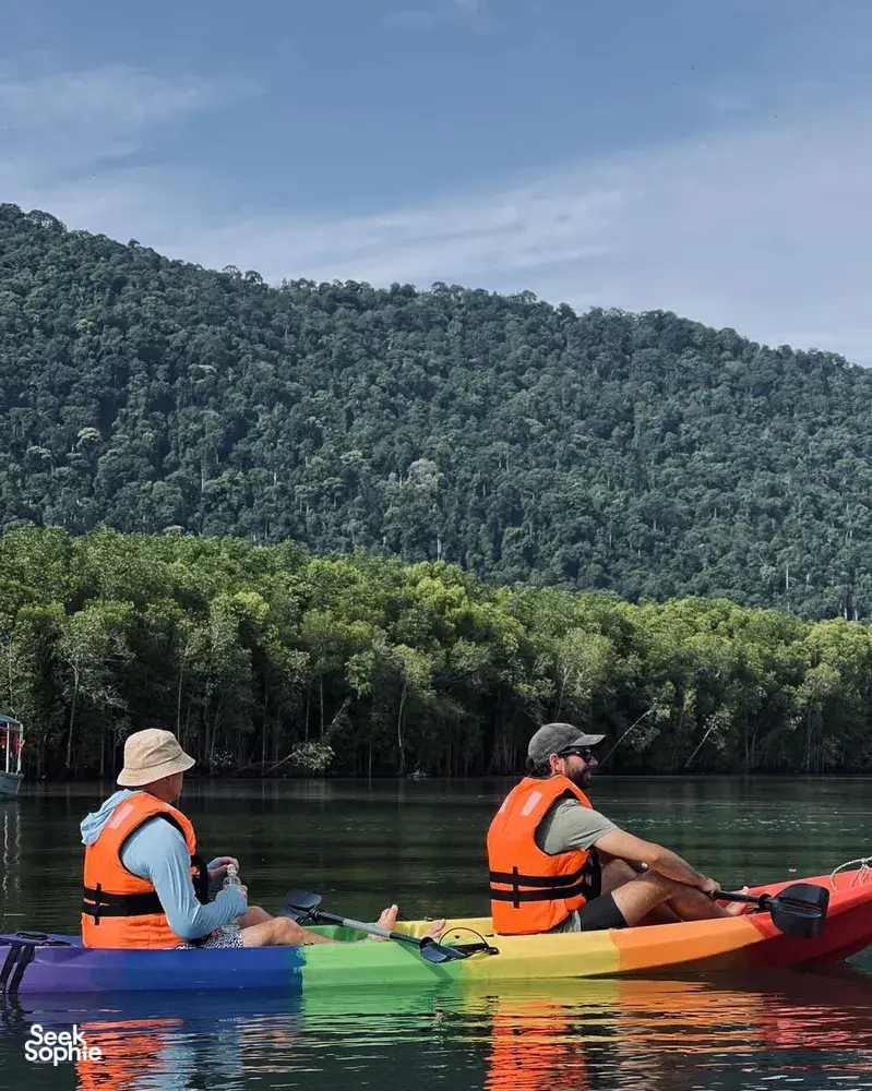 Kayakers in orange life jackets paddle along a mangrove-lined river in Kubang Badak, Langkawi, under forested hills