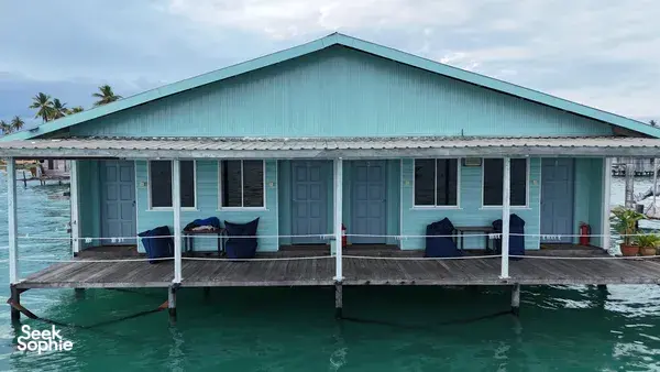 Banglo air kayu berwarna hijau kebiruan di atas tiang di Semporna, dengan tempat duduk di anjung hadapan dan laluan pejalan kaki di atas laut yang tenang