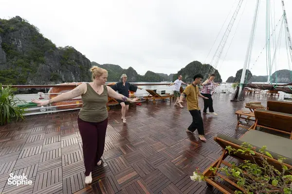 Group stretching on the deck of a Cat Ba boutique cruise, with limestone karst cliffs under an overcast sky at the bay