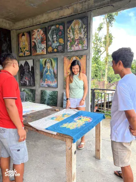 In the Five Arts Studio gallery in Keliki village, a craftsperson and visitors view batik art on a work table in Bali, Ubud