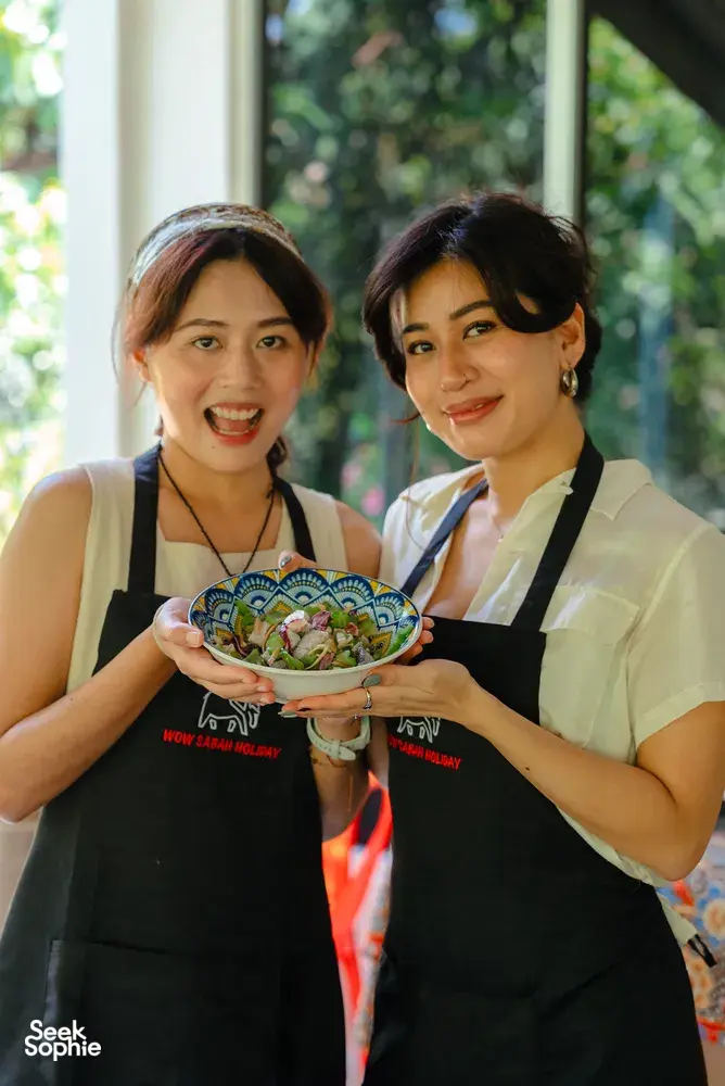 Two Indigenous Sabahan women in aprons holding a colourful bowl of salad during a local cooking experience in Kota Kinabalu, Sabah