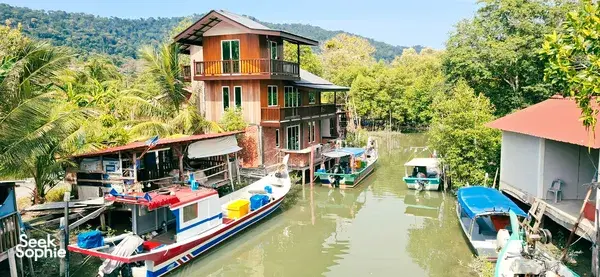 Row of tied boats beside a narrow Langkawi canal, with riverside houses, palm trees and green hills in view