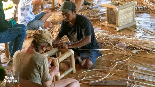 Indonesia bamboo woodworking class: an instructor and participants tie and weave bamboo strands, with tools and bindings on the floor