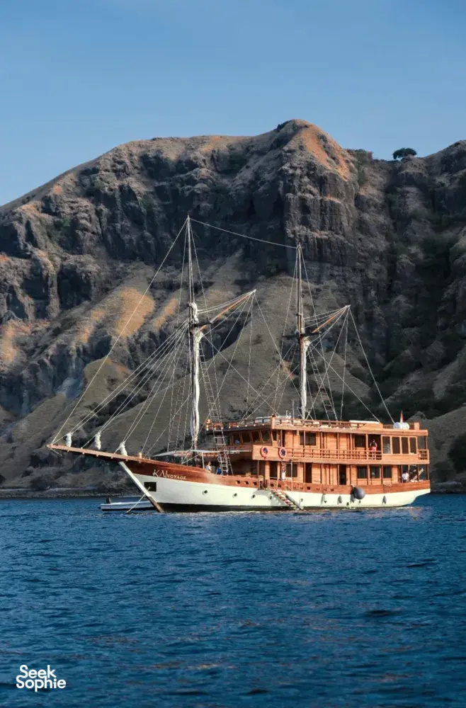 Sailing phinisi passing rugged volcanic hills in calm blue water, for a 3-day shared Komodo cruise from Labuan Bajo